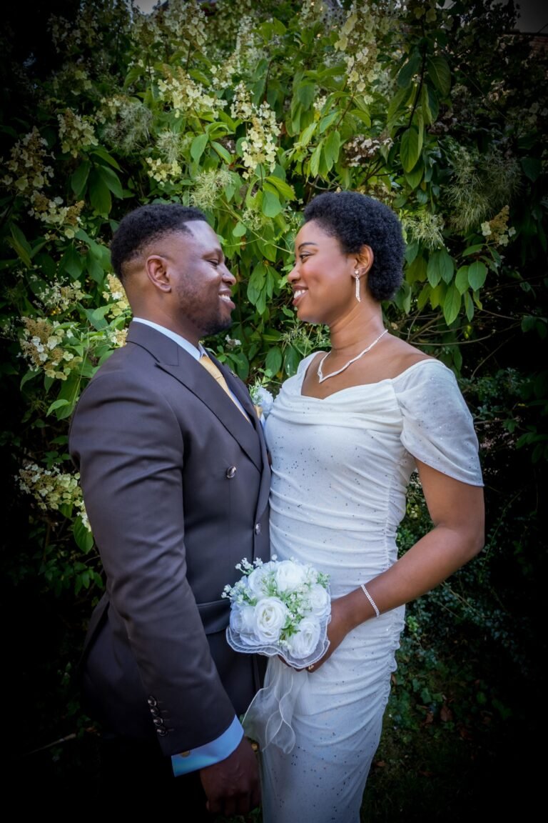 Bride and groom facing each other in front of green foliage, the bride holding a white bouquet
