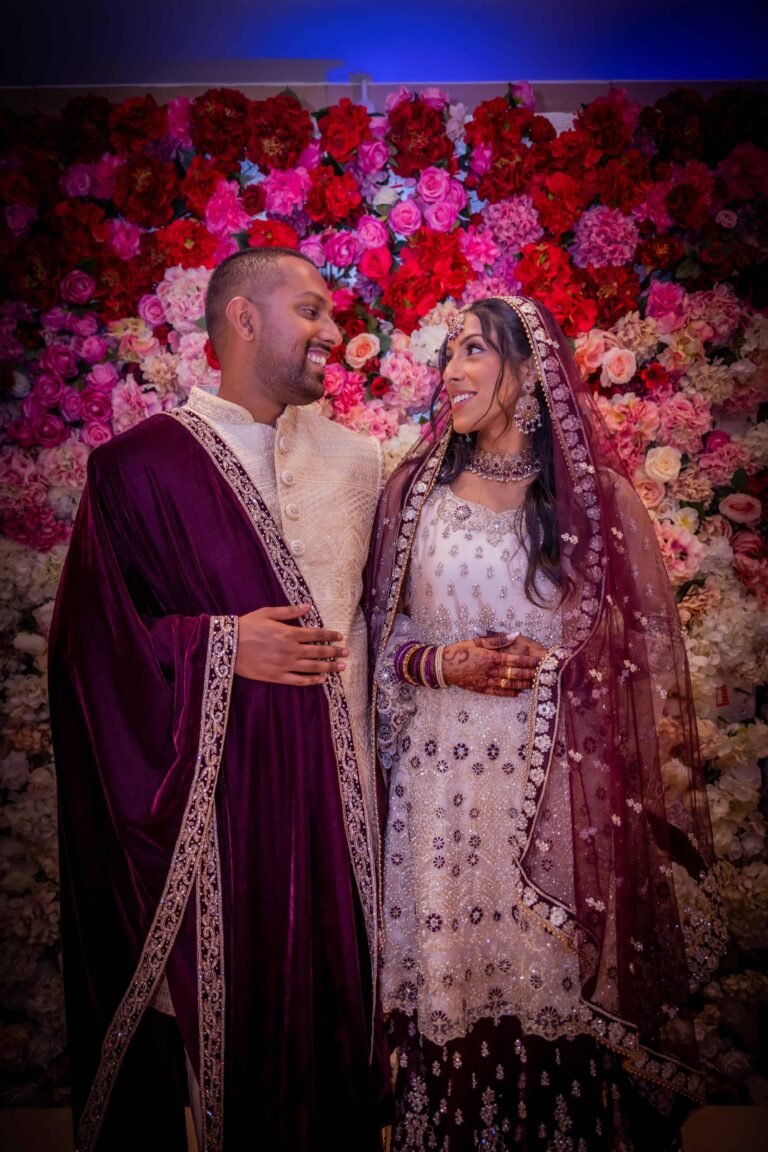 Couple before a pink and red rose wall, the groom in a maroon sherwani