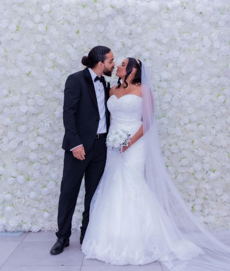 Bride and groom nose to nose at a white flower wall, the bride holding a bouquet