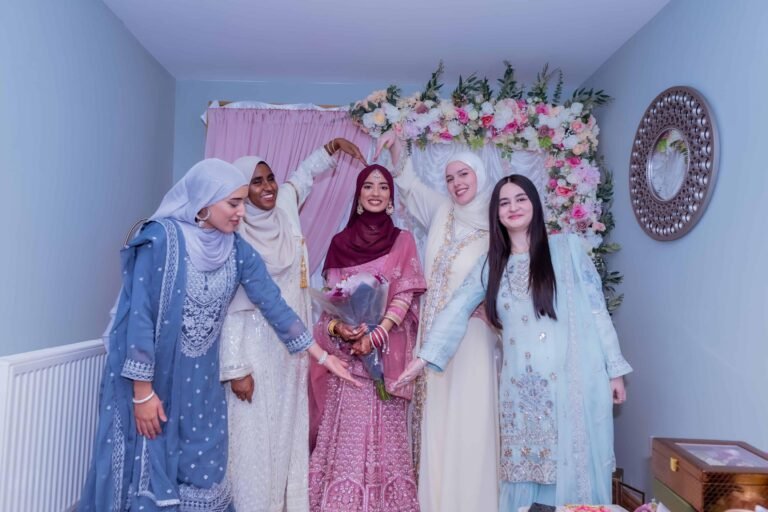 Bride with female guests in pastel outfits and hijabs beneath a floral arch