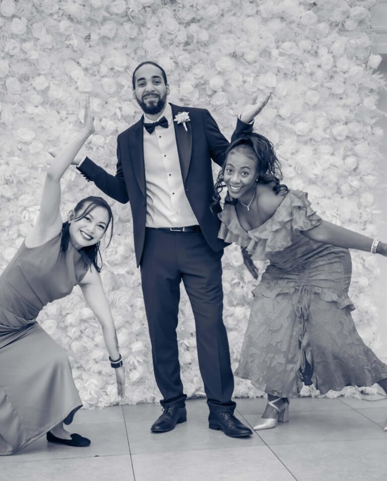 Playful black and white shot of a groom and two bridesmaids posing at a flower wall