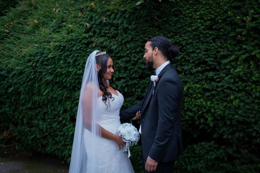 Bride and groom facing each other in front of a tall green hedge on their wedding day